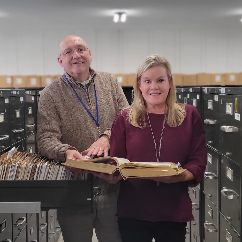 Two records staff in a file-room aisle reviewing a bound ledger amid rows of lateral filing cabinets.